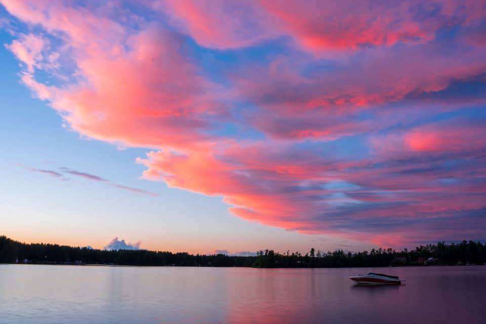 A calm lake with a boat, surrounded by a forest and a colorful sky.