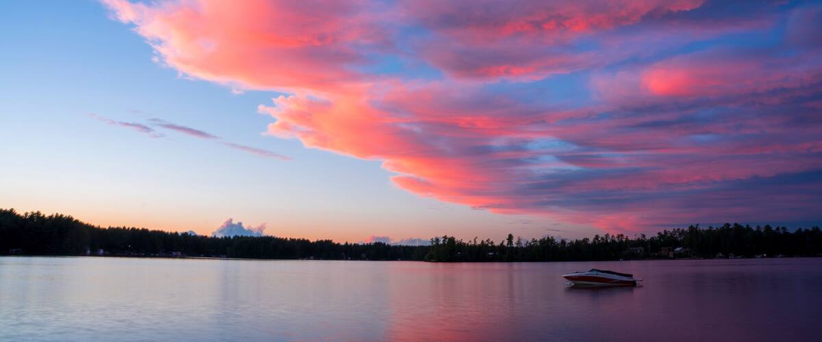 A calm lake with a boat, surrounded by a forest and a colorful sky.