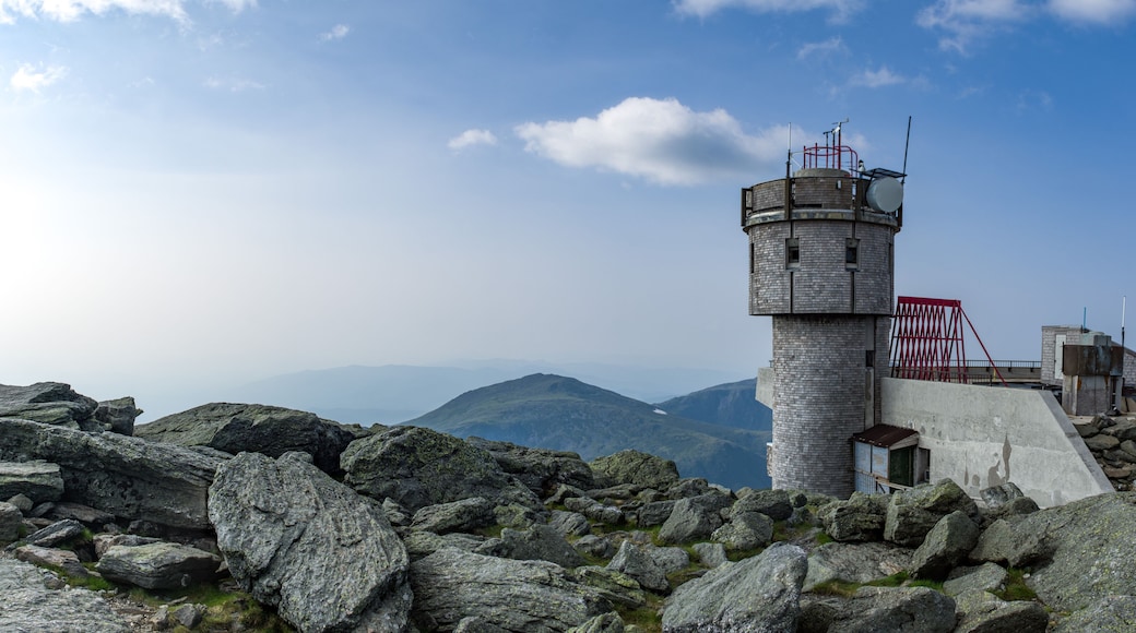 Un faro de piedra sobre un promontorio rocoso, cielo despejado y montañas al fondo.