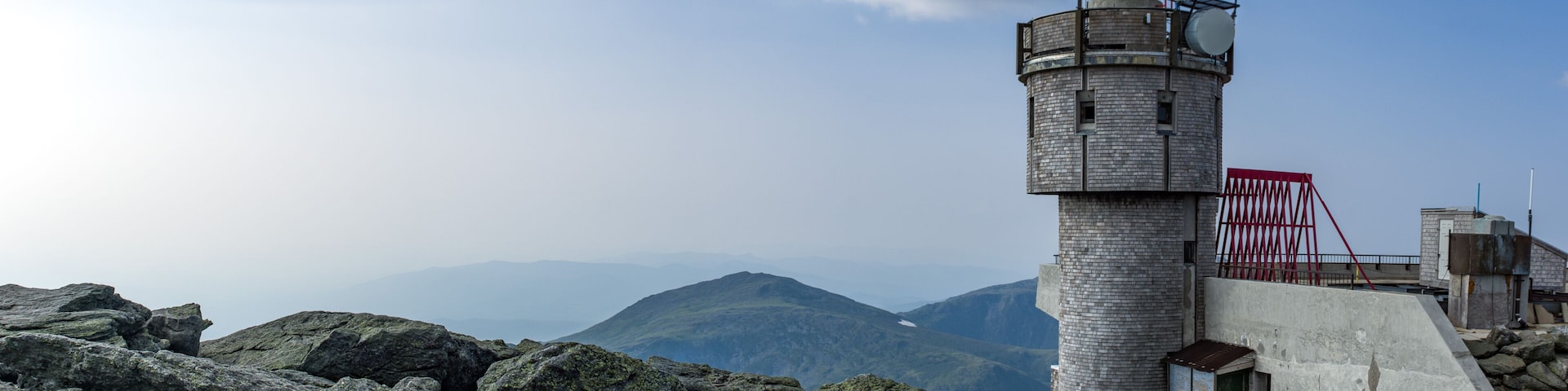 Um farol de pedra numa falésia rochosa, com céu claro e montanhas ao fundo.