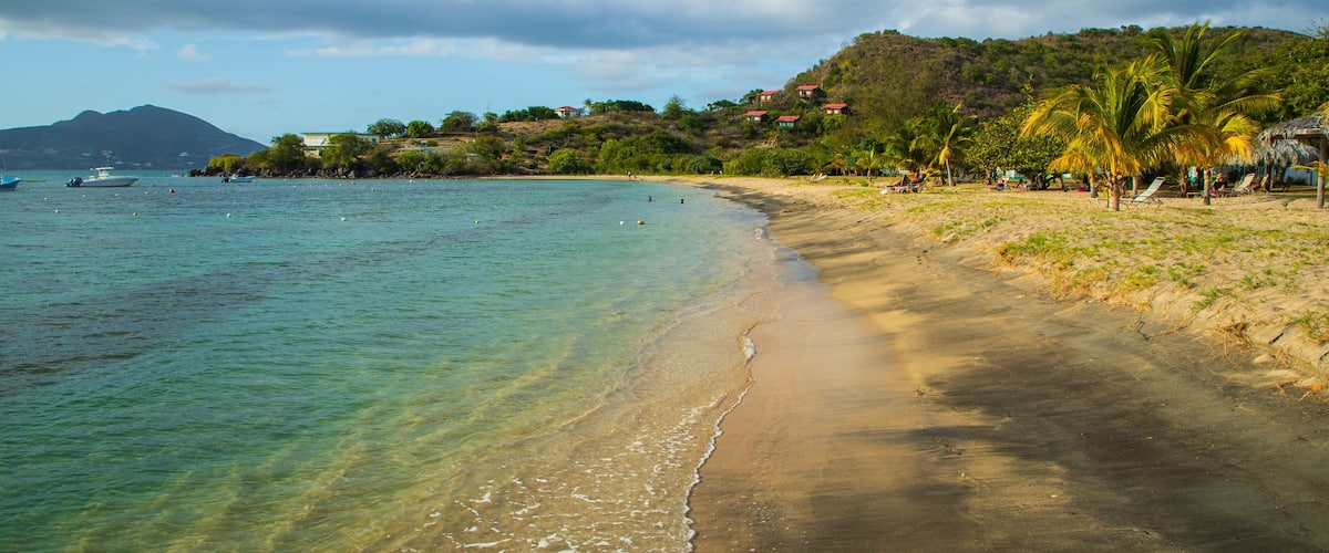 A beach with clear turquoise water, a sandy shore, and a backdrop of hills with houses.