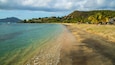 A beach with clear turquoise water, a sandy shore, and a backdrop of hills with houses.