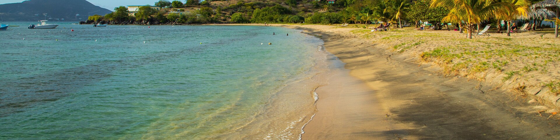Una playa con agua turquesa cristalina, una orilla arenosa y colinas al fondo con casas.