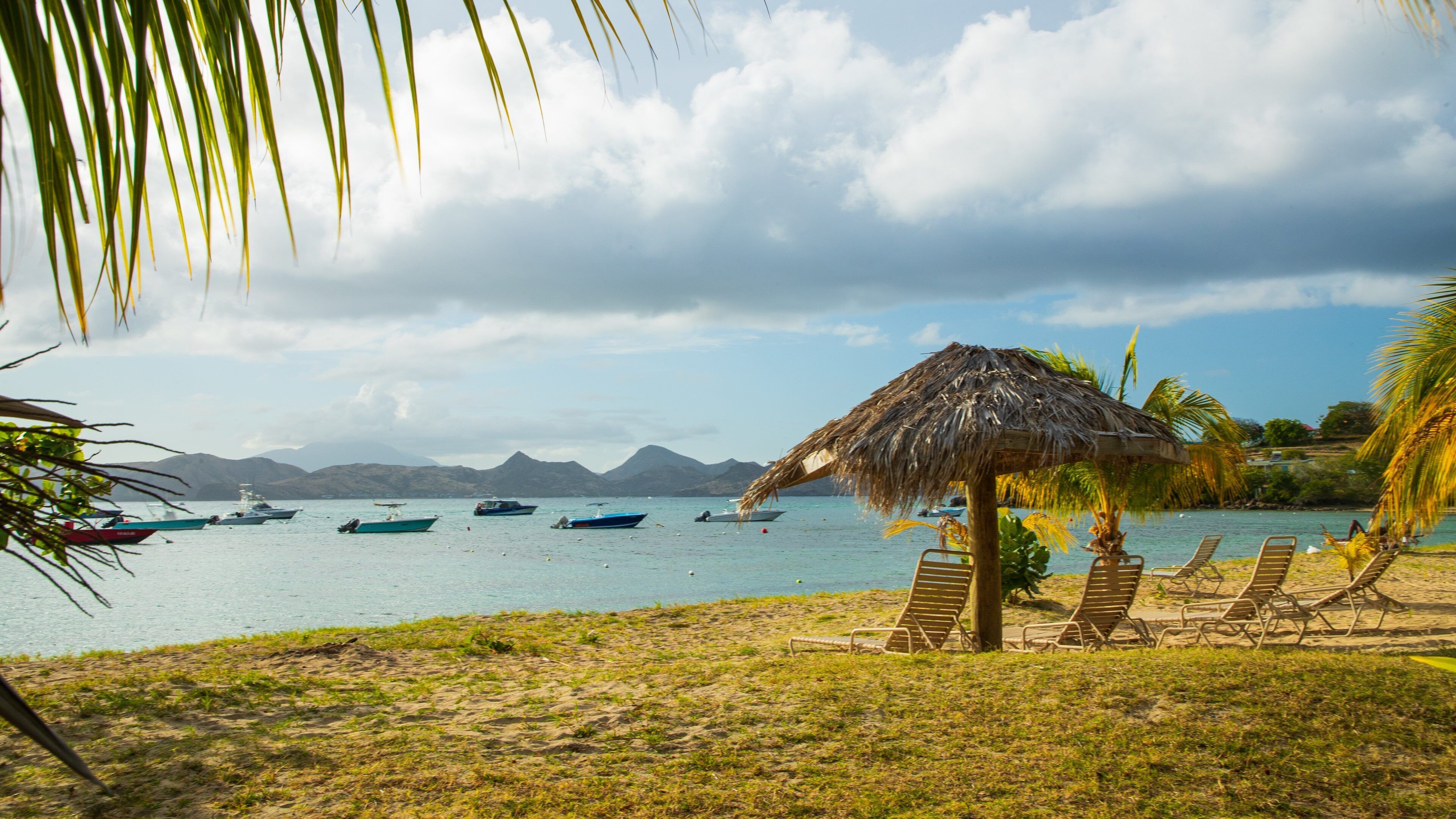 A beach with a thatched hut, lounge chairs, and boats on calm water with mountains in the background.