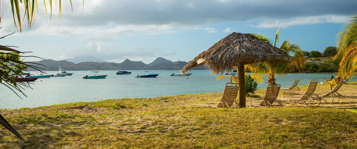A beach with a thatched hut, lounge chairs, and boats on calm water with mountains in the background.