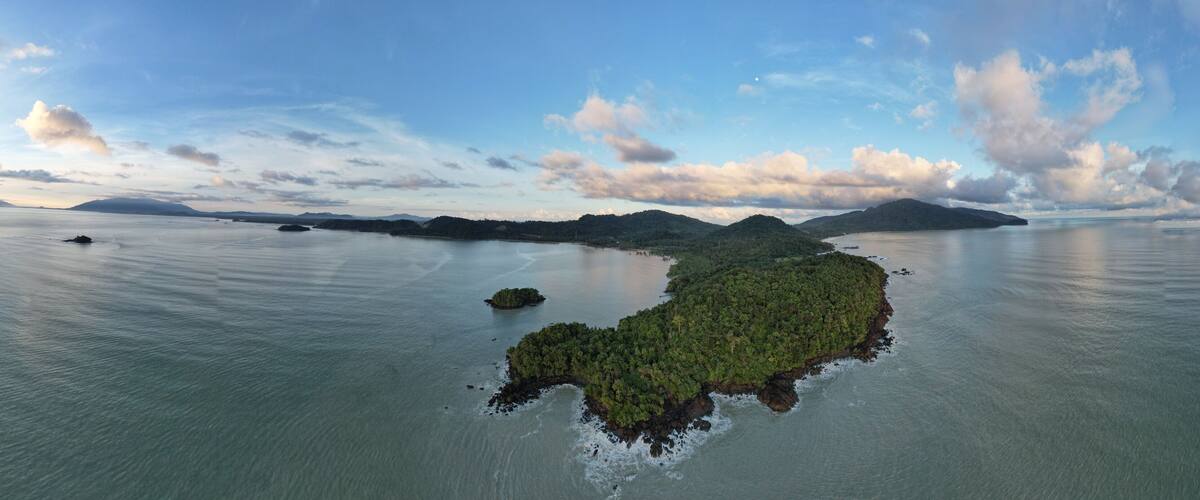 A coastal landscape with a large island surrounded by calm waters, a distant mountain range, and a partly cloudy sky.