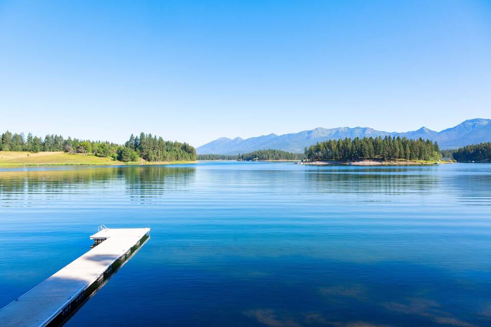Een rustig meer met een steiger die het water in reikt, omgeven door dichte bossen en verafgelegen bergen, onder een heldere blauwe lucht.