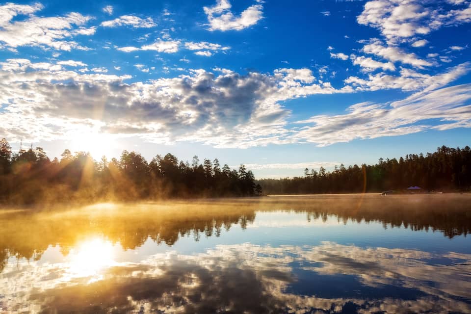 Un llac tranquil amb reflexos d'arbres i núvols, envoltat per un bosc.