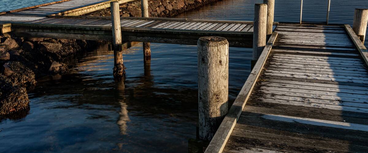 A wooden pier extending into calm water, with a stone wall on one side and a grassy area with a building in the background.