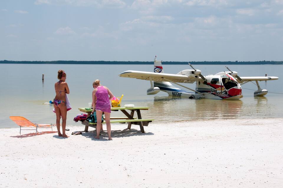 A beach with two people standing near a picnic table and a small seaplane on the water.