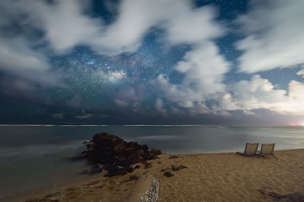 A beach with two lounge chairs, a rocky outcrop, and a starry night sky.