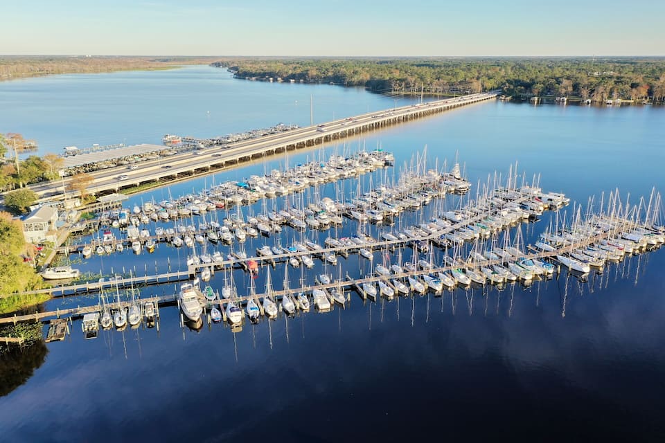 A marina with numerous boats docked along piers, surrounded by calm water and a distant bridge.