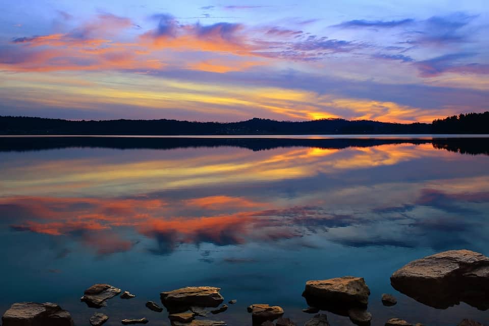 A calm lake with a sunset reflection, surrounded by rocks and a forested hill in the background.