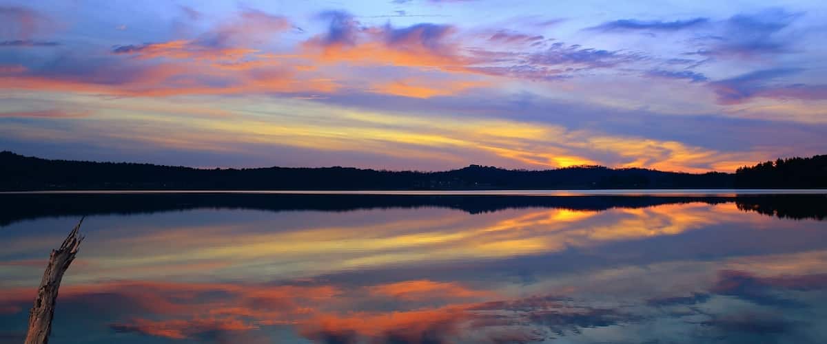 A calm lake with a sunset reflection, surrounded by rocks and a forested hill in the background.