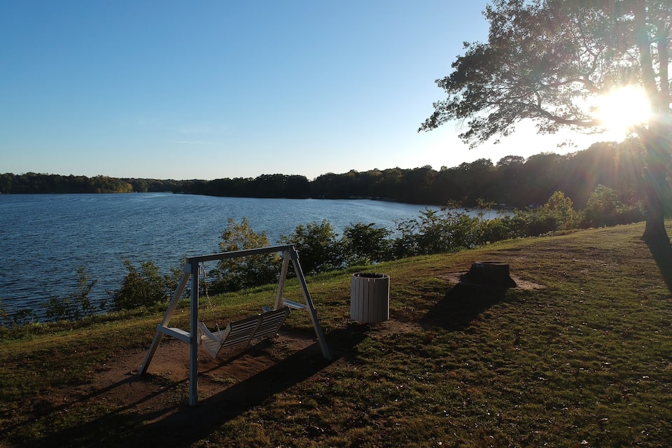 A lakeside park with a swing set, a trash can, and a tree casting a shadow.