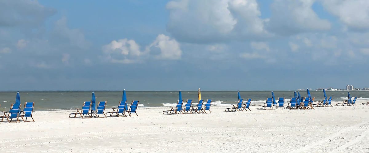 A beach with rows of blue beach chairs and umbrellas, a sandy shore, and the ocean in the background.