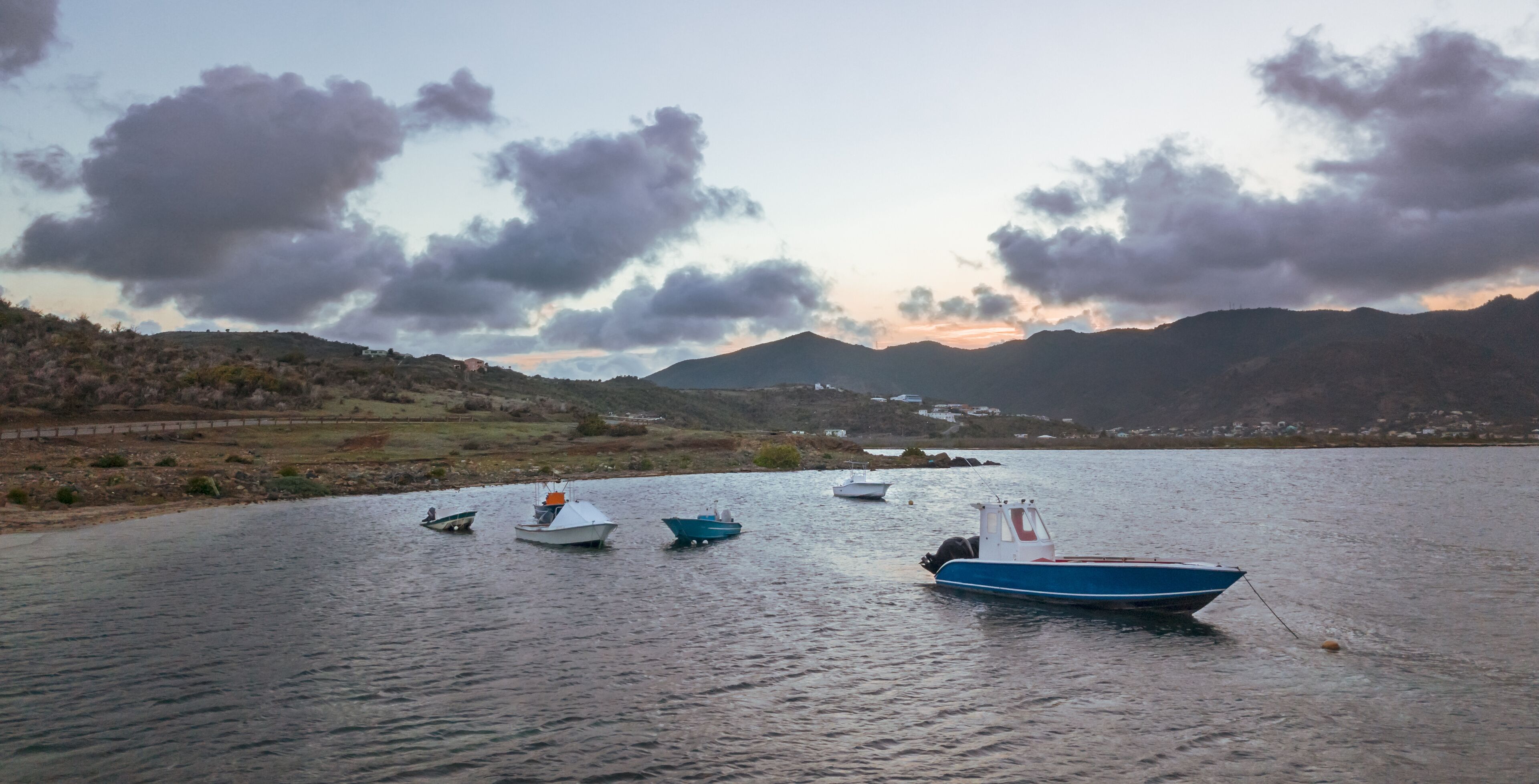 A calm lake with several boats anchored, surrounded by hills and a cloudy sky.