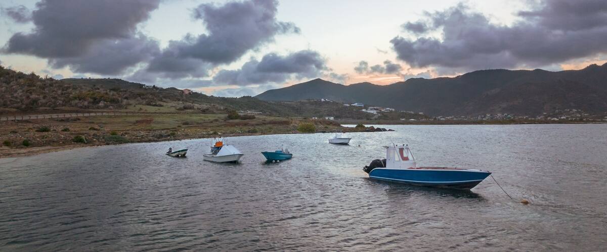 A calm lake with several boats anchored, surrounded by hills and a cloudy sky.