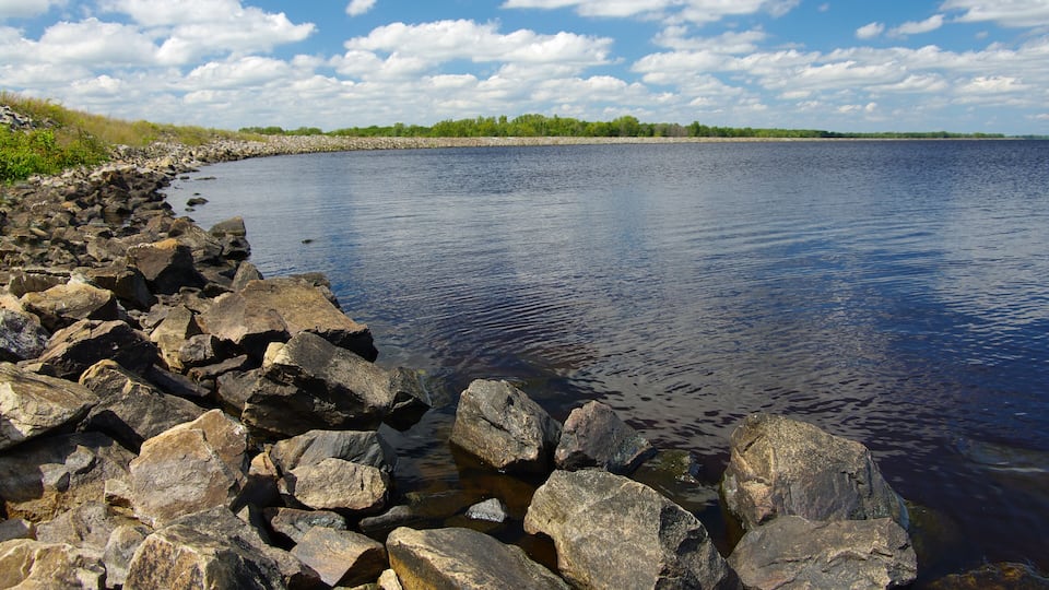 A rocky shoreline with a calm lake and a distant tree line under a partly cloudy sky.