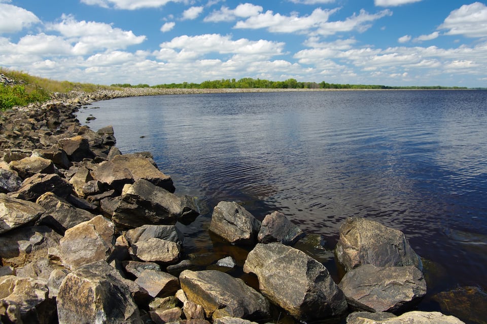 A rocky shoreline with a calm lake and a distant tree line under a partly cloudy sky.