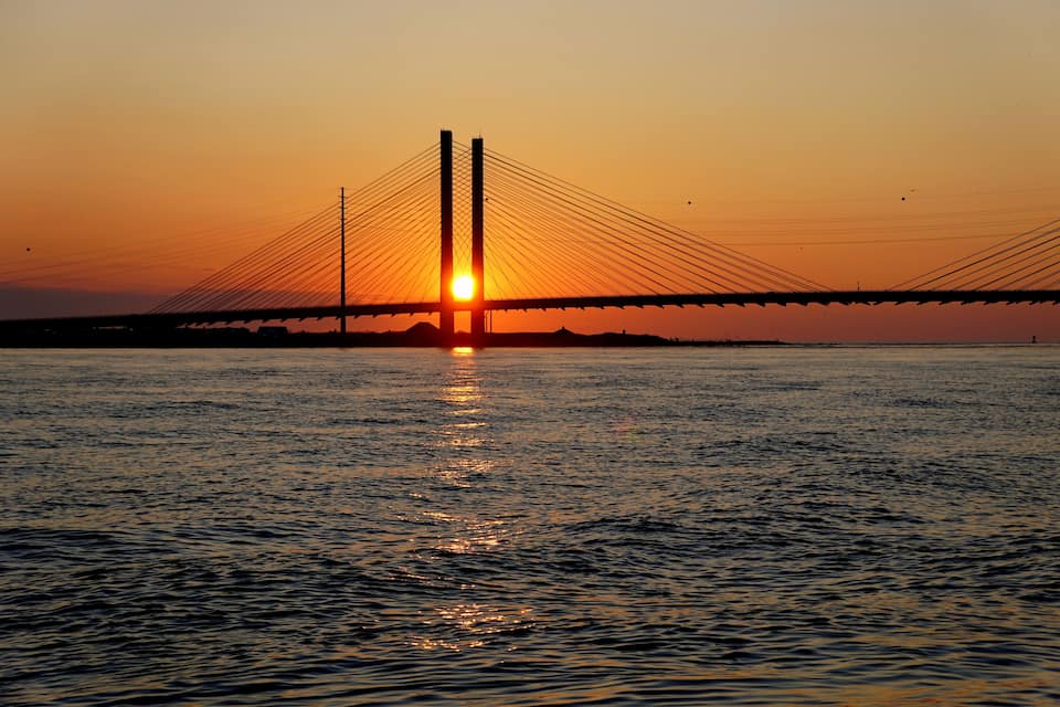 A suspension bridge at sunset with the sun reflecting on the water.