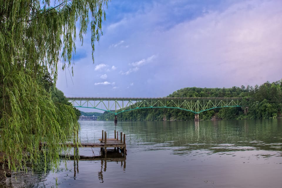 A river with a wooden dock and a green bridge.