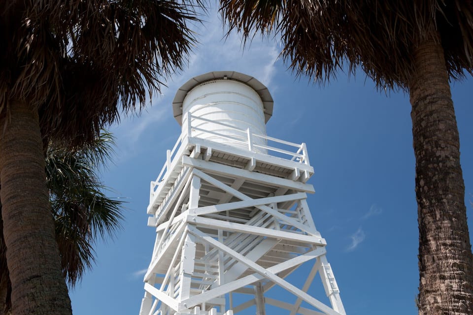 Een witte stalen vuurtoren met een wenteltrap, omringd door palmbomen en een heldere blauwe lucht.