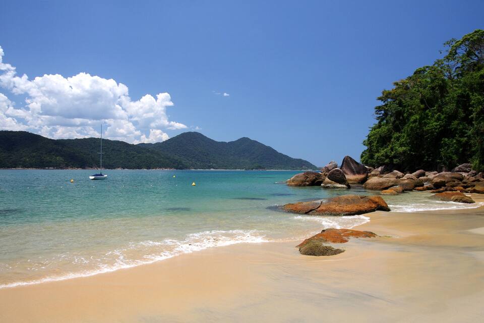 A beach with clear turquoise water, a sailboat, and large rocks.