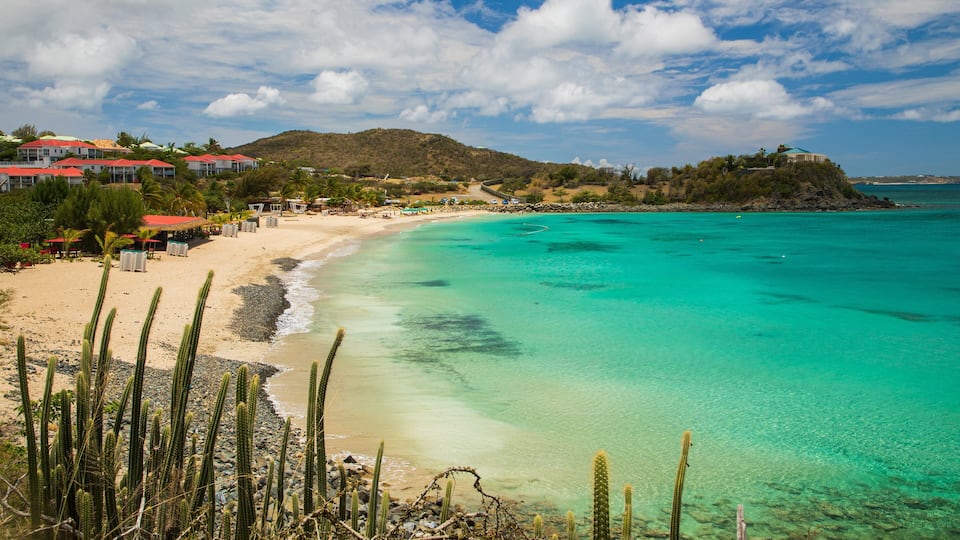 A beach with clear turquoise water, a sandy shore, and a backdrop of buildings and hills.