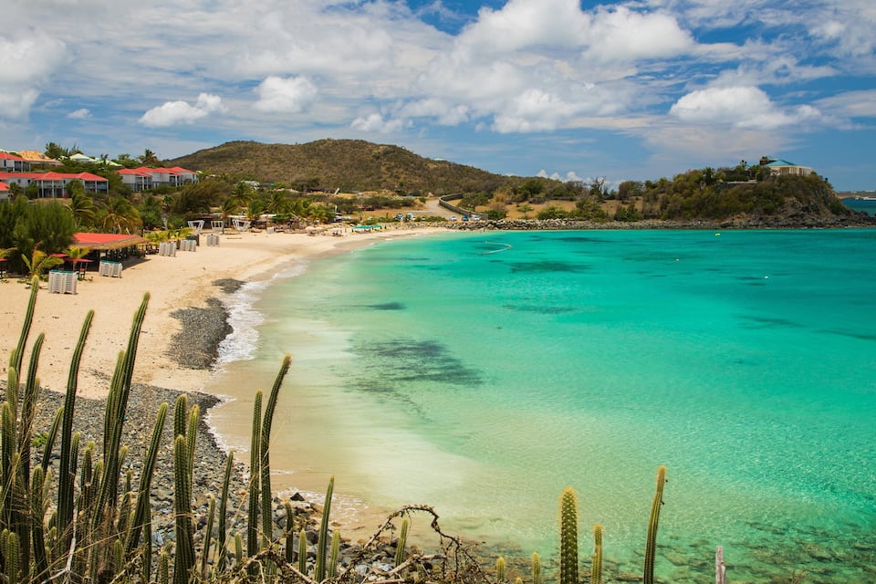Ein Strand mit klarem türkisblauem Wasser, einem sandigen Ufer und im Hintergrund Gebäuden und Hügeln.