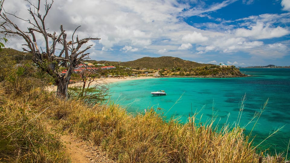 A coastal landscape with turquoise waters, a boat, and a beach with buildings in the background.