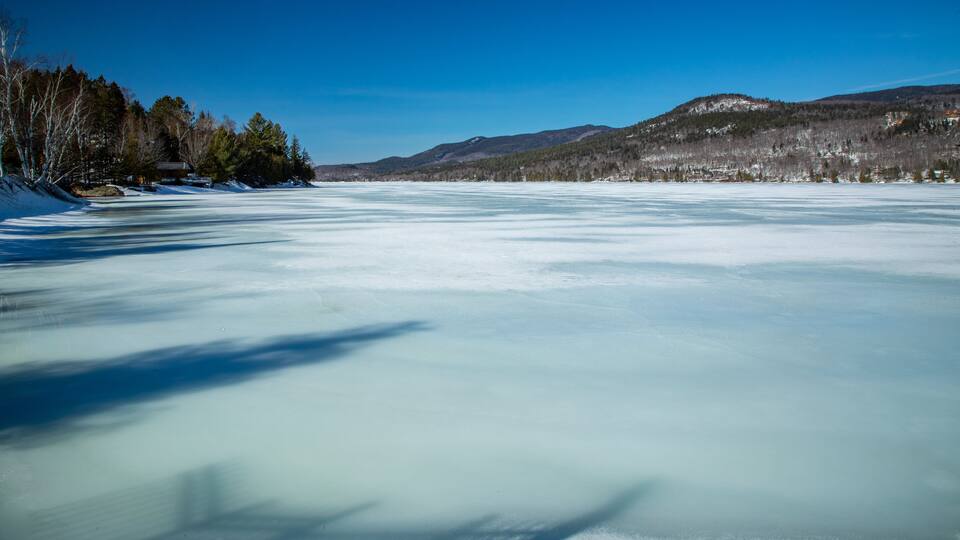 A frozen lake with snow-covered trees and mountains in the background.