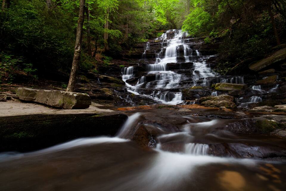 A multi-tiered waterfall in a forested area with rocky terrain and lush greenery.