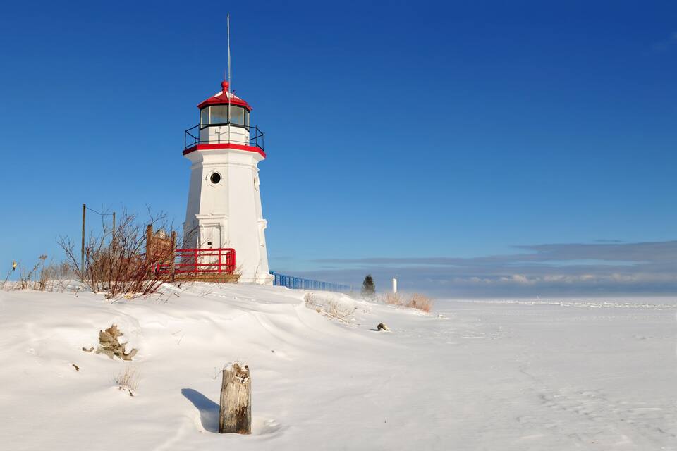 A white lighthouse with a red top and balcony, surrounded by snow and a clear blue sky.