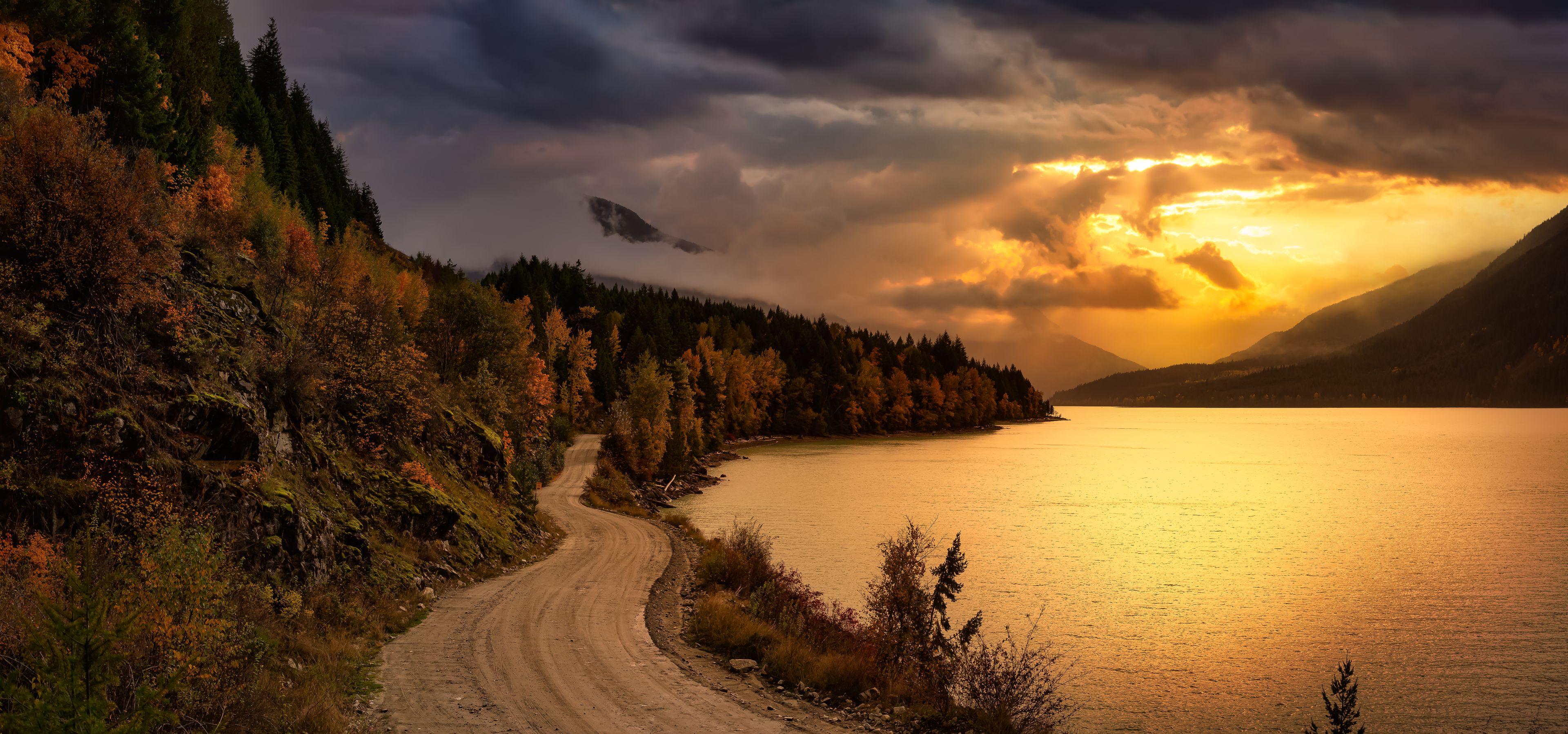 A dirt road winding through a forested area beside a calm lake with mountains in the background and a dramatic sunset sky.