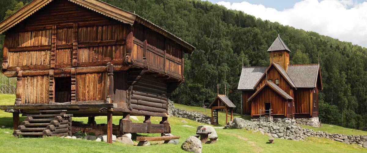 A wooden log cabin with a steeply pitched roof, surrounded by a stone wall and a stone staircase.