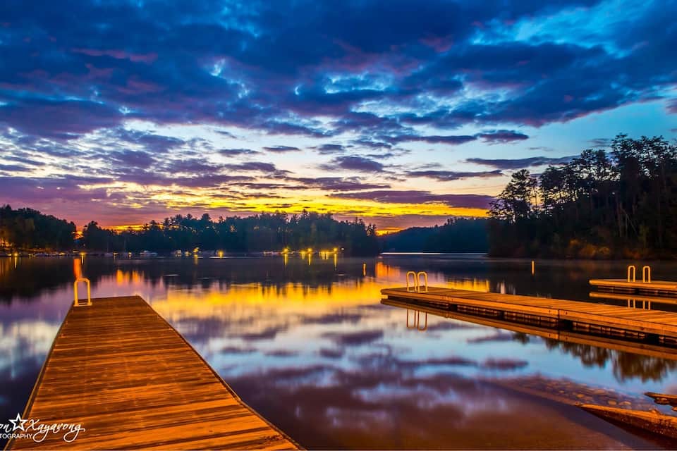 A wooden pier extending into a calm lake at sunset with a colorful sky reflected on the water.