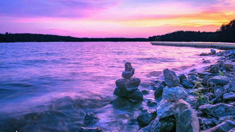 A sunset over a lake with a stack of rocks in the foreground and a pier in the background.