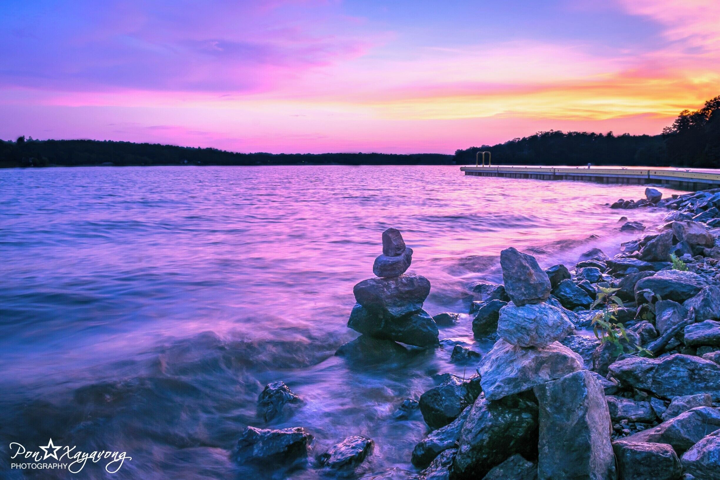 A sunset over a lake with a stack of rocks in the foreground and a pier in the background.