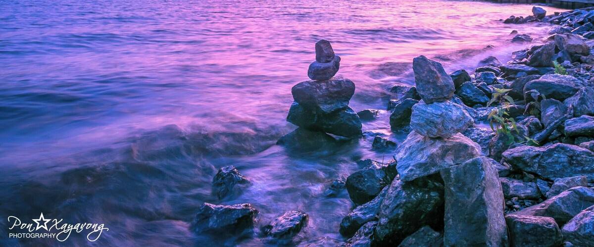 A sunset over a lake with a stack of rocks in the foreground and a pier in the background.