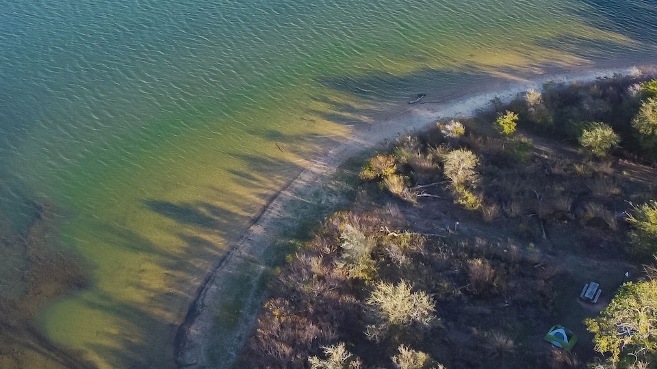 An aerial view of a coastal area with a sandy beach, a clear water body, and a forested area.