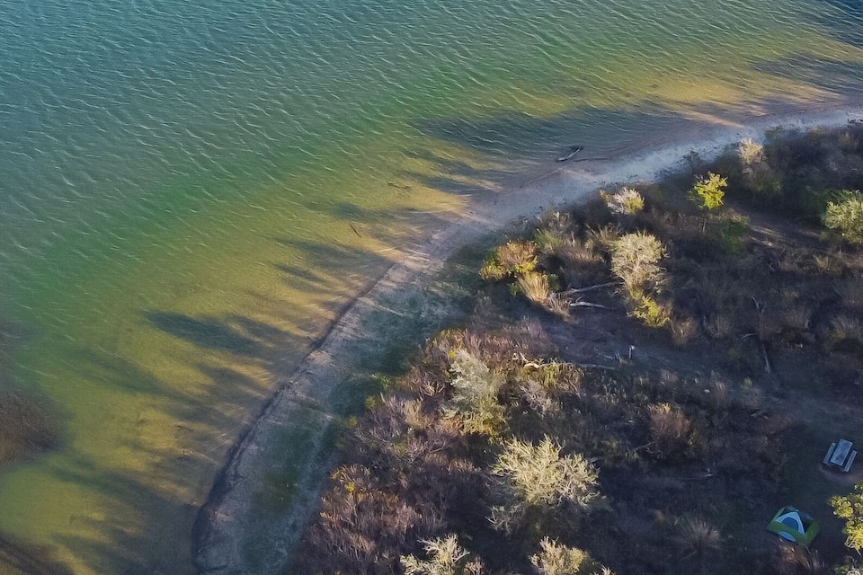 An aerial view of a coastal area with a sandy beach, a clear water body, and a forested area.