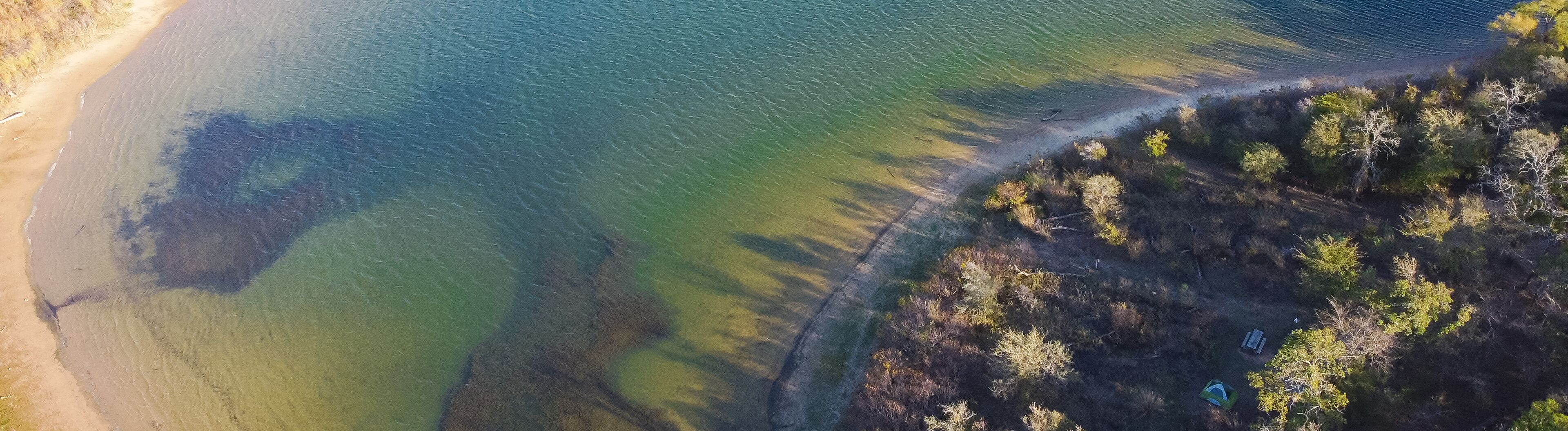 An aerial view of a coastal area with a sandy beach, a clear water body, and a forested area.