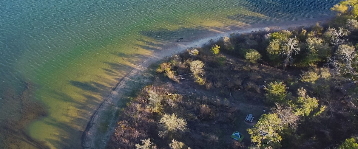 An aerial view of a coastal area with a sandy beach, a clear water body, and a forested area.