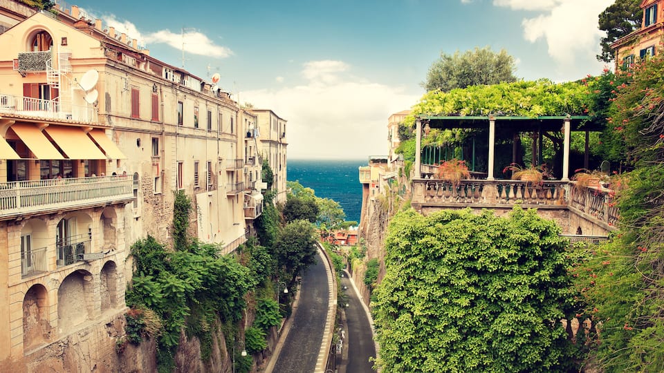 A narrow street flanked by historic buildings with balconies overlooking a coastal view.