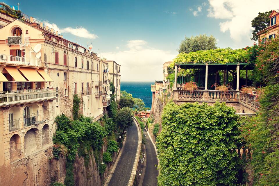 A narrow street flanked by historic buildings with balconies overlooking a coastal view.