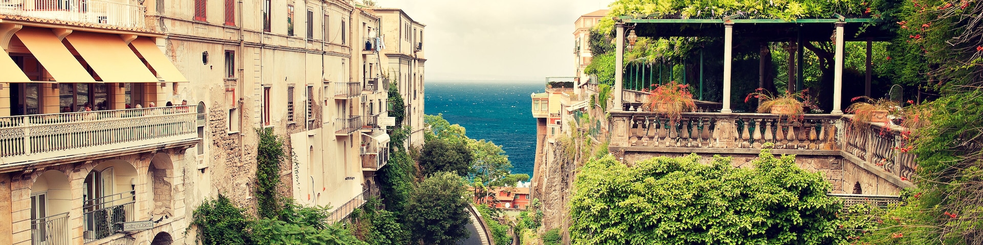 A narrow street flanked by historic buildings with balconies overlooking a coastal view.