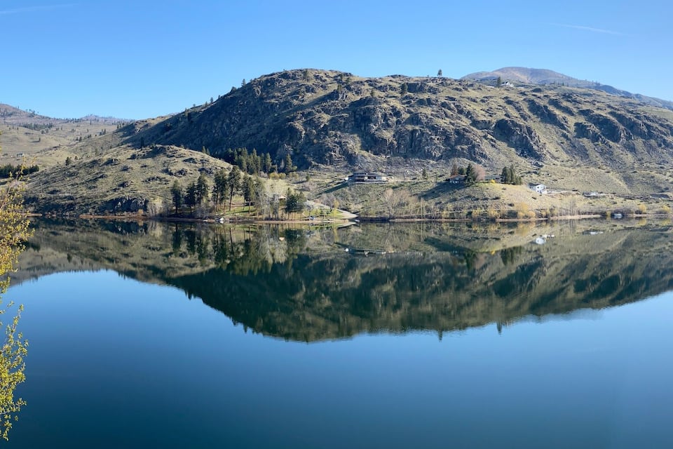 Um lago calmo refletindo uma paisagem montanhosa com vegetação escassa e um céu claro.