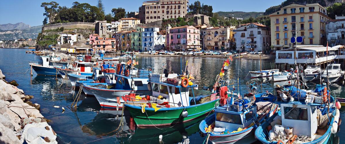 A harbor with colorful boats docked along the shore, surrounded by buildings and a mountainous backdrop.