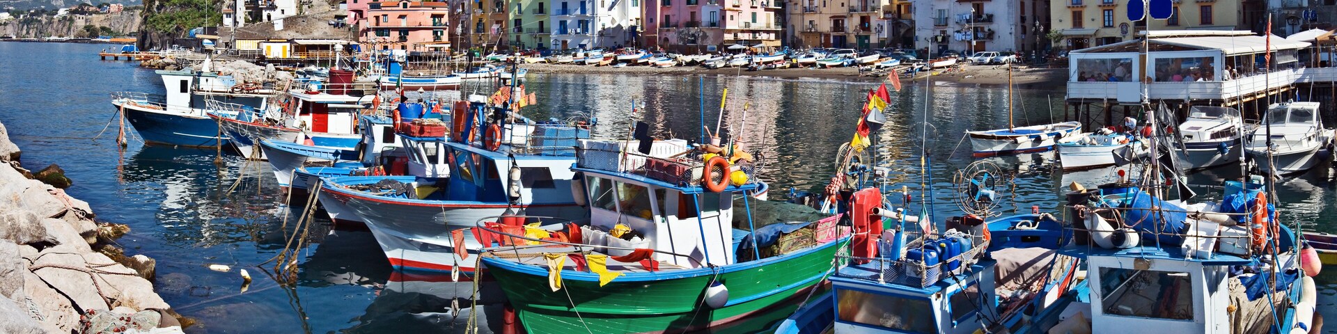 A harbor with colorful boats docked along the shore, surrounded by buildings and a mountainous backdrop.